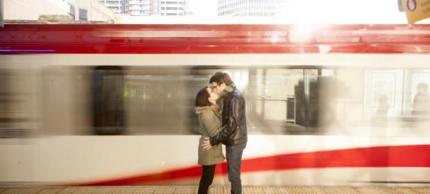 A man and woman stand kissing while a gray and red train passes behind them.