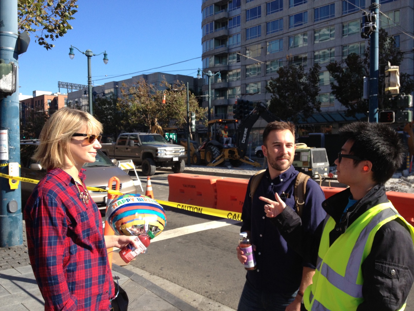 A man and woman stop to get directions and information from one of the project ambassadors in a black jacket and yellow safety vest. King Street and construction equipment are in the background.