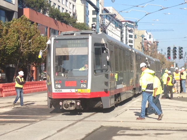 Construction and project staff in safety gear stand on either side of a Muni train as the operator tests the new tracks..