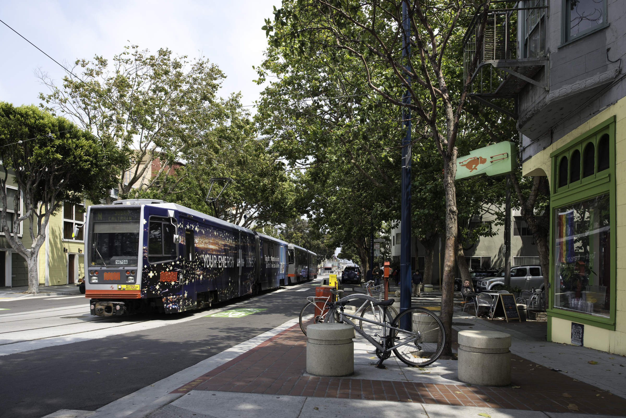 N Line light rail train with a dark blue advertising wrap travels east on a tree-lined Duboce Street past the Duboce Park Cafe. A bicycle is parked on the sidewalk. It and the street furniture are in the foreground.
