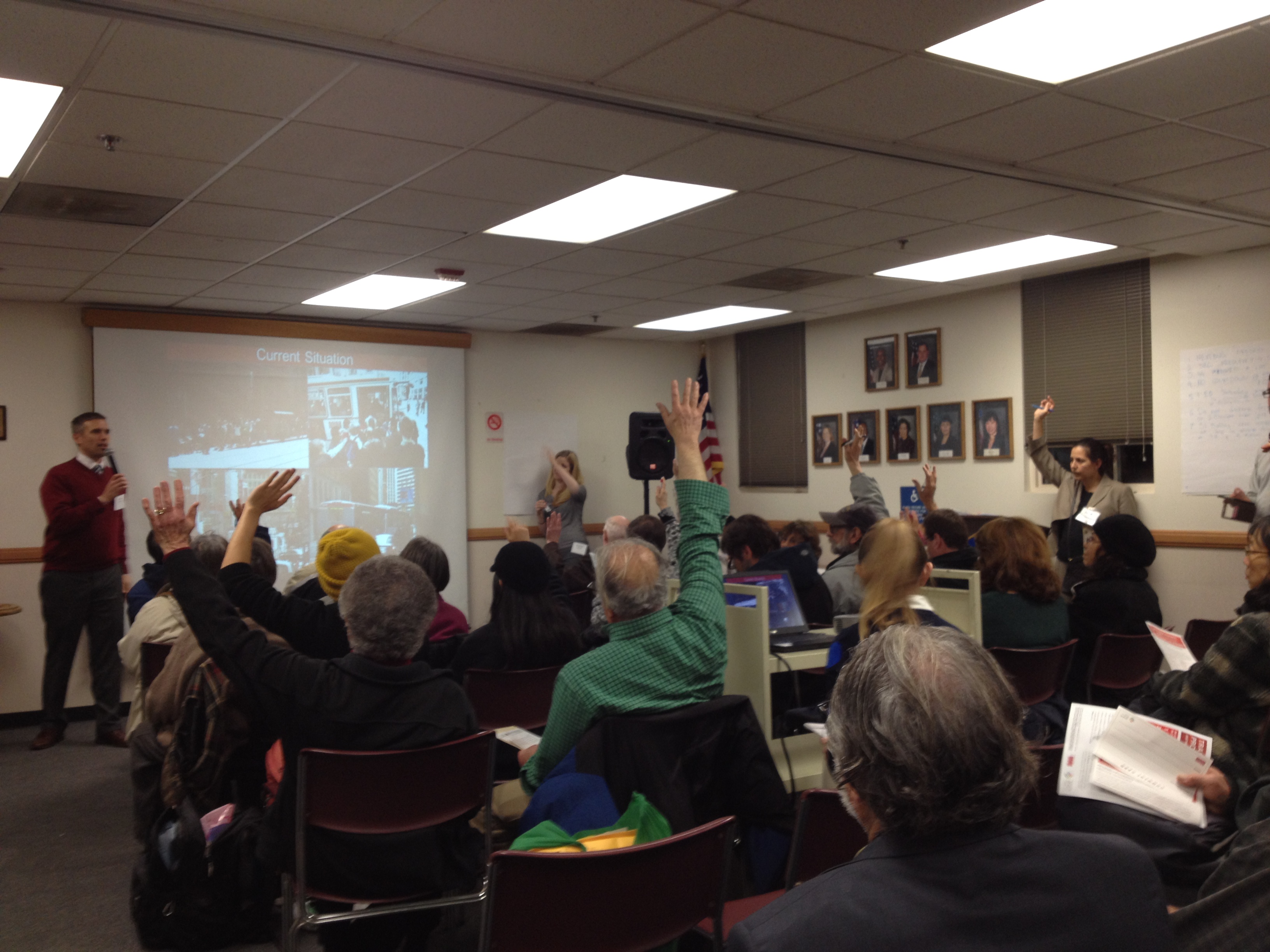 Seated members of the public raise their hands as a man stands with a microphone at the front of the room 