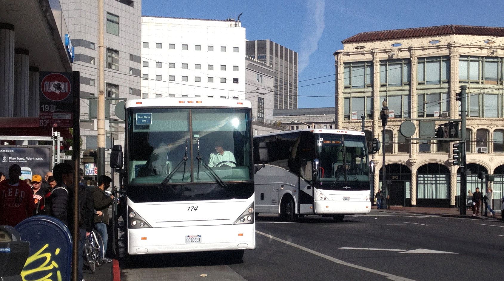 Two white buses board riders along a busy sidewalk.