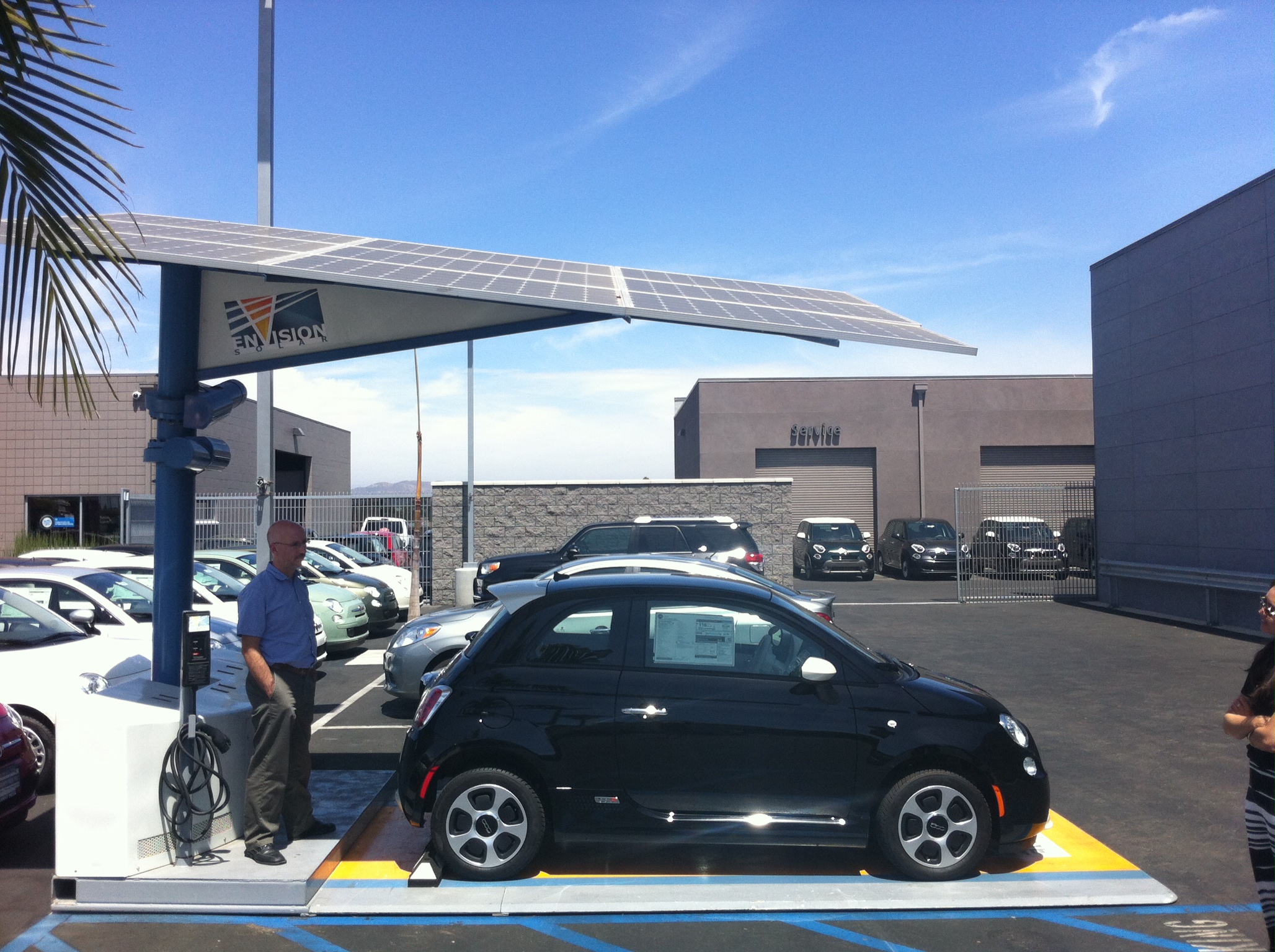 A man standing next to a small black car underneath a large solar panel in a sunny parking lot next to a palm tree.