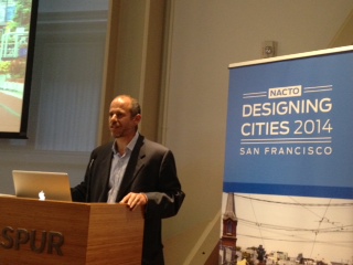Ed Reiskin stands at a lecturn speaking to the NACTO Designing Cities 2014 conference. The conference blue sign with white text is behind him.