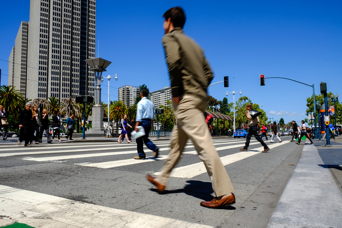 Man in beige sportscoat and slacks walks east with other pedestrians across The Embarcadero with SF high-rises and blue sky in the background