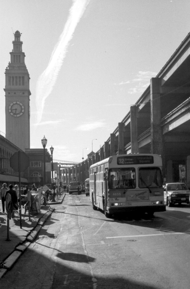 Black and White photograph of the 32 Embarcadero Muni bus driving north on the Embarcadero with the shuttered skyway on the right and the Ferry Building clock tower on the left. 