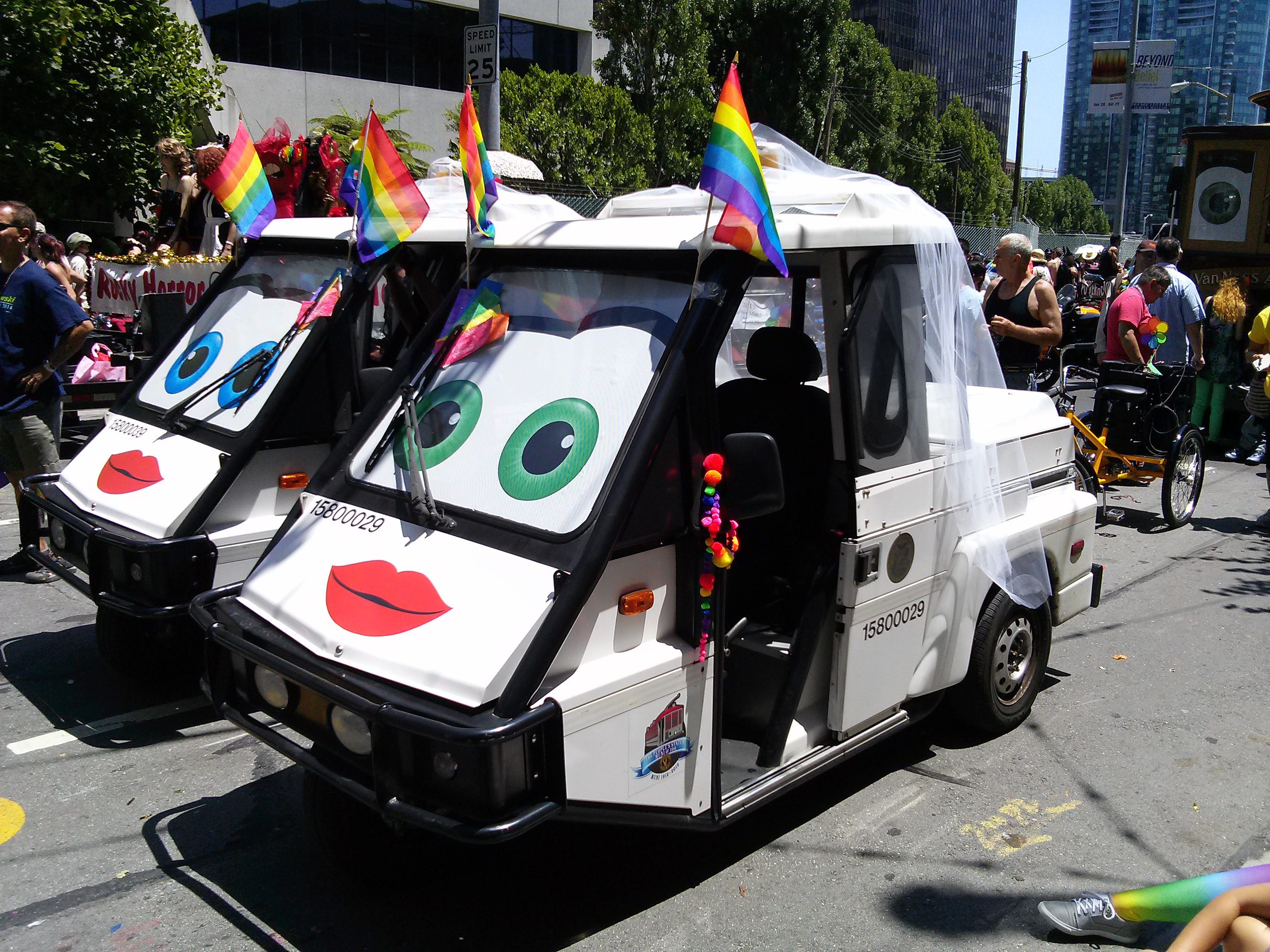 Two parking control white, three-wheeled vehicles decorated with anthropomorphic eyes and lips sport wedding veils and rainbow flags.