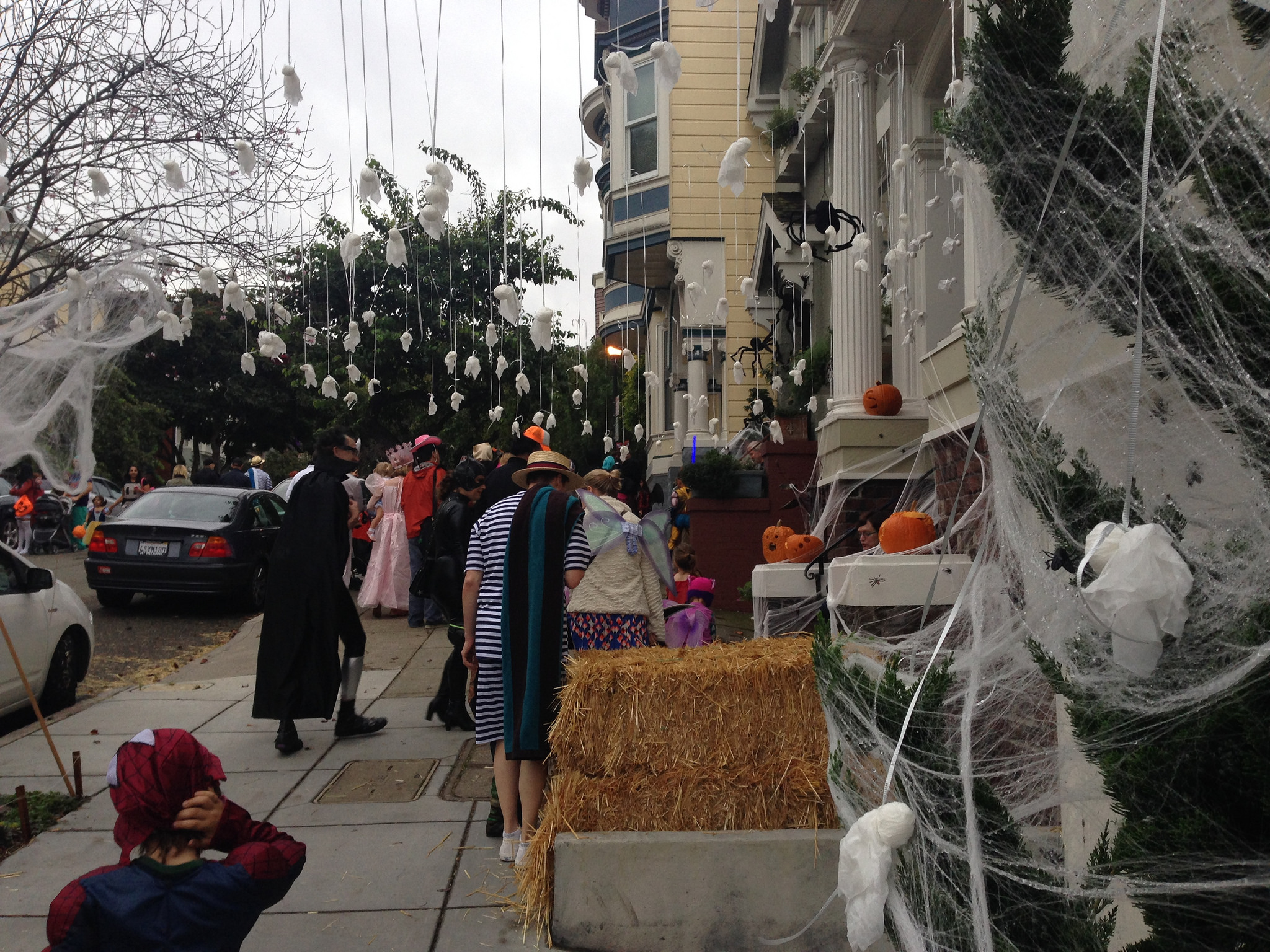 Neighborhood street with adults and children in costumes, bales of hay, ghosts and spiderwebs decorations