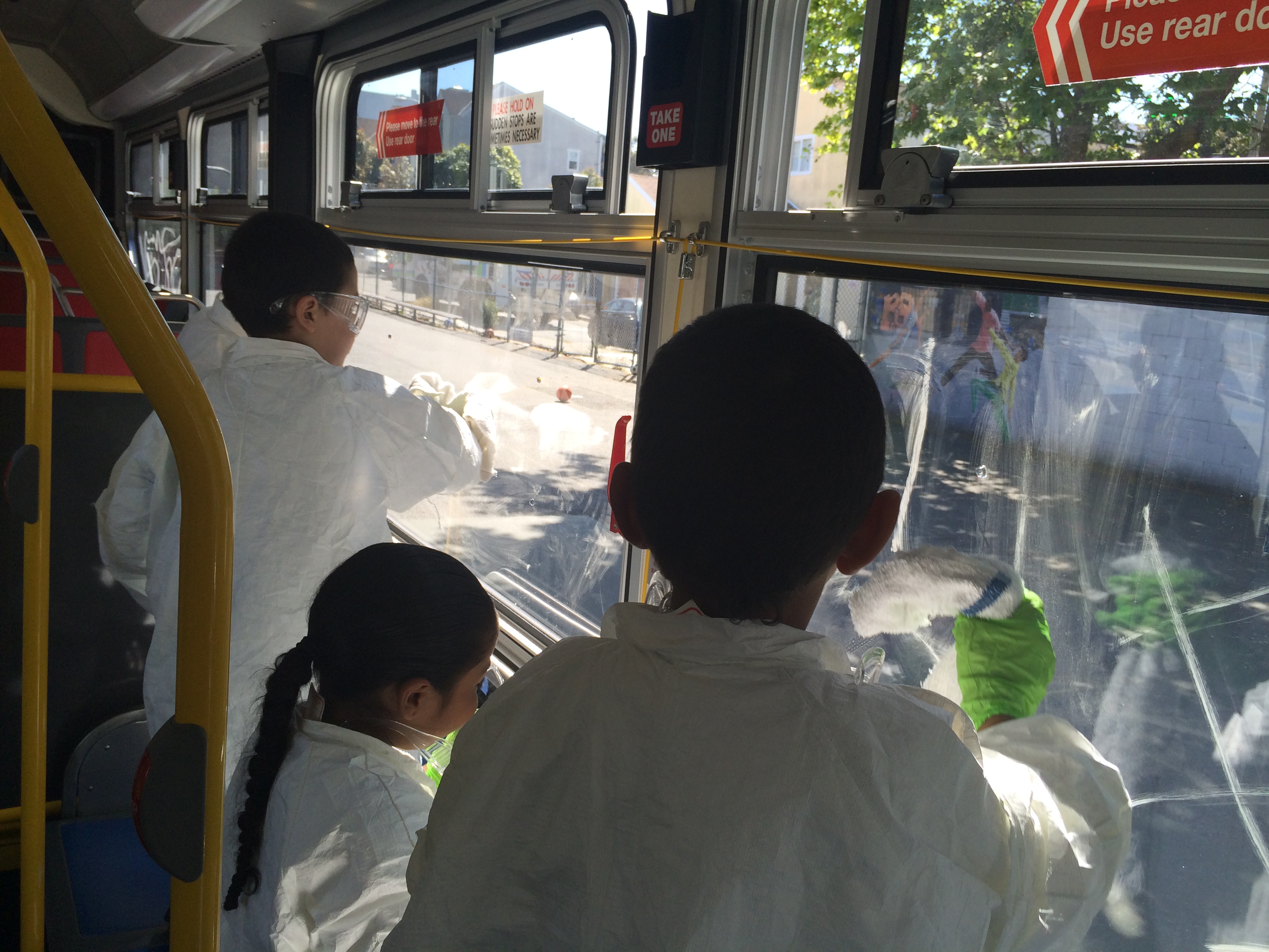 Three young children in white smocks and green gloves clean a window inside of a Muni bus