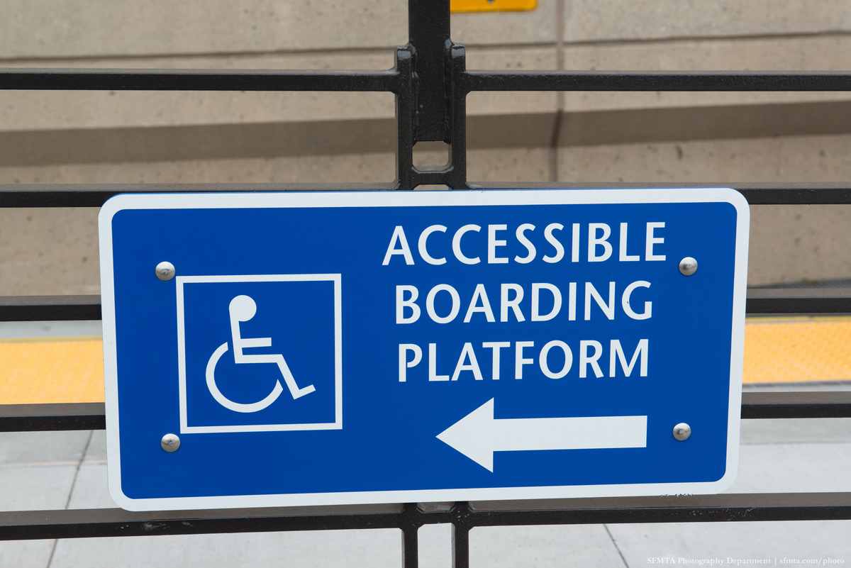 White text, "Accessbile Boarding Platform" with a white arrow below it, next to a white International Symbol of Access on a blue field affixed to a railing near a boarding platform.