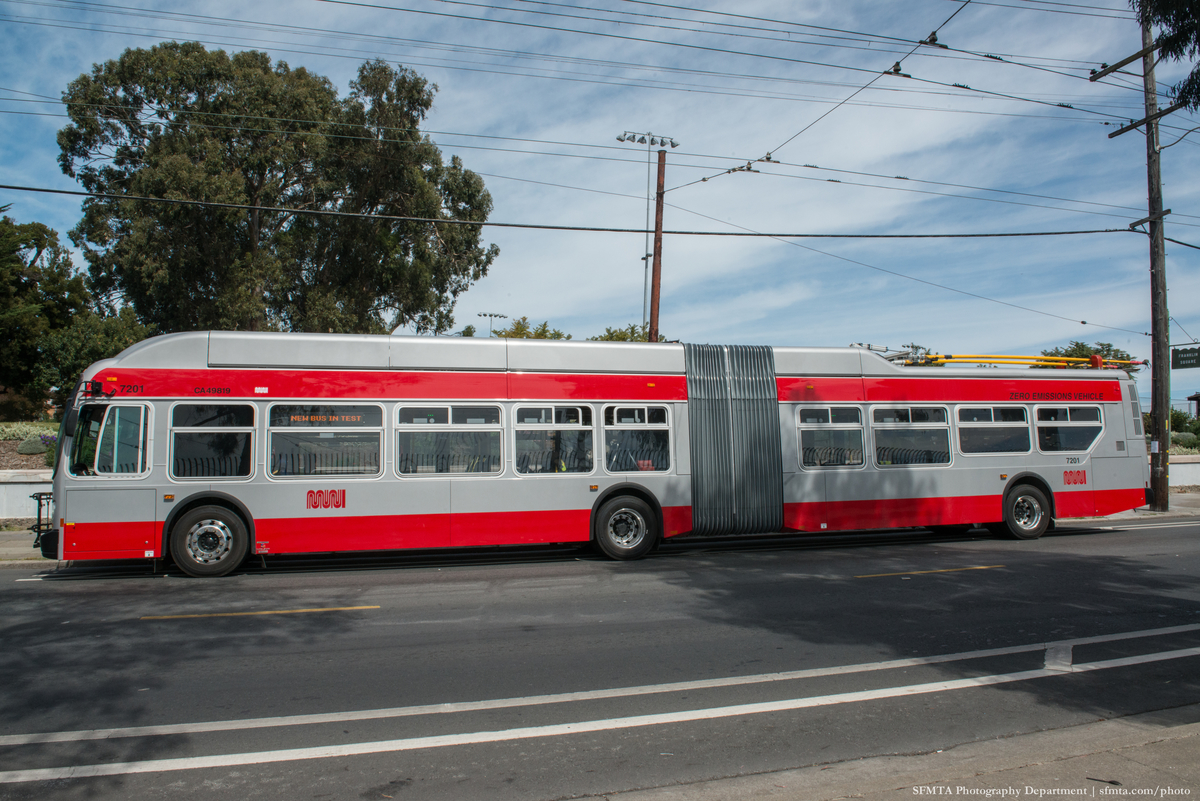 Side view of new 60-foot trolley bus in the current gray and red Muni livery with the bright yellow polls lying on top. Tall trees and overhead wires in the background.