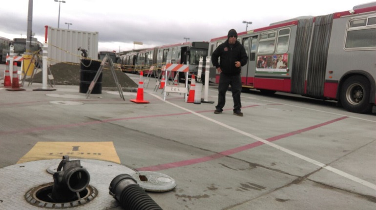 A hose sits next to a connector for an underground gasoline storage tank in a Muni yard with a man in black cold-weather gear and two gray and red Muni buses in the background.