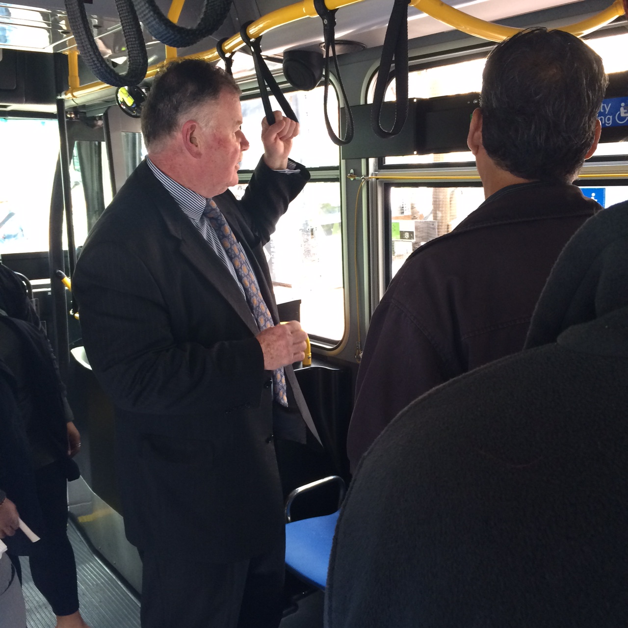 A man in a suit stands on a new Muni bus with two other men standing to his right while he explains features of the bus.