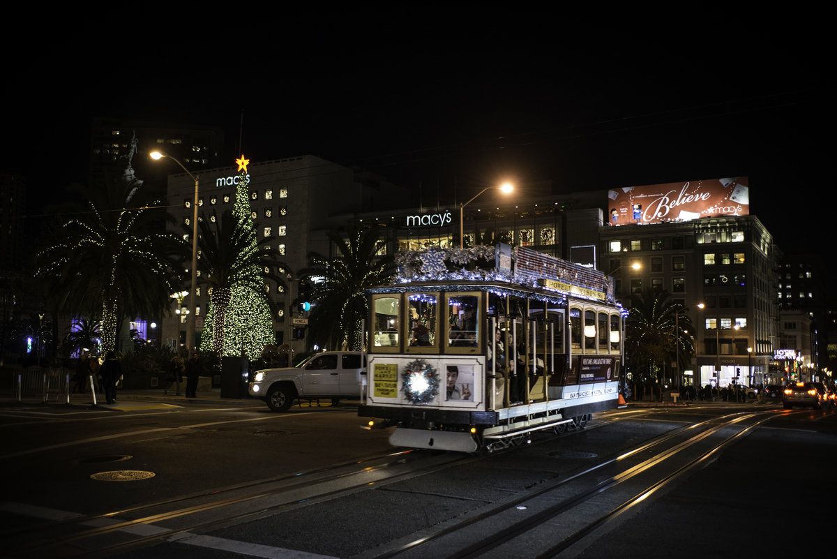 A cable car with lights and a wreath heads north on Powell Street at night past Union Square with Macy's and the Christmas Tree in the background.