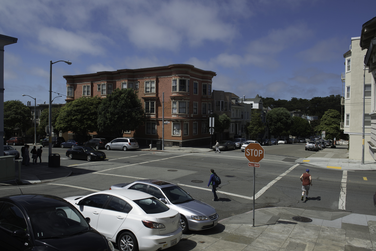 High-angle shot of a San Francisco intersection with cars parked in lower left corner and pedestrians walking through white-striped crosswalks.