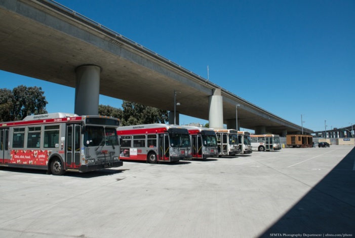 Red and gray modern Muni buses lined up under a freeway overpass
