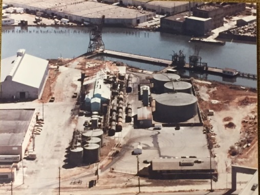 Aerial photograph of industrial buildings and tanks with a wide creek running along the top of the photograph.