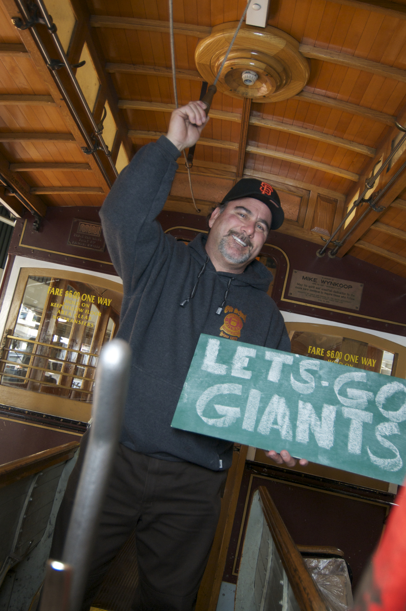 Man in Giant's baseball hat and cable car sweatshirt stands on a cable car, ringing the bell with a sign reading "Let's Go Giants!)