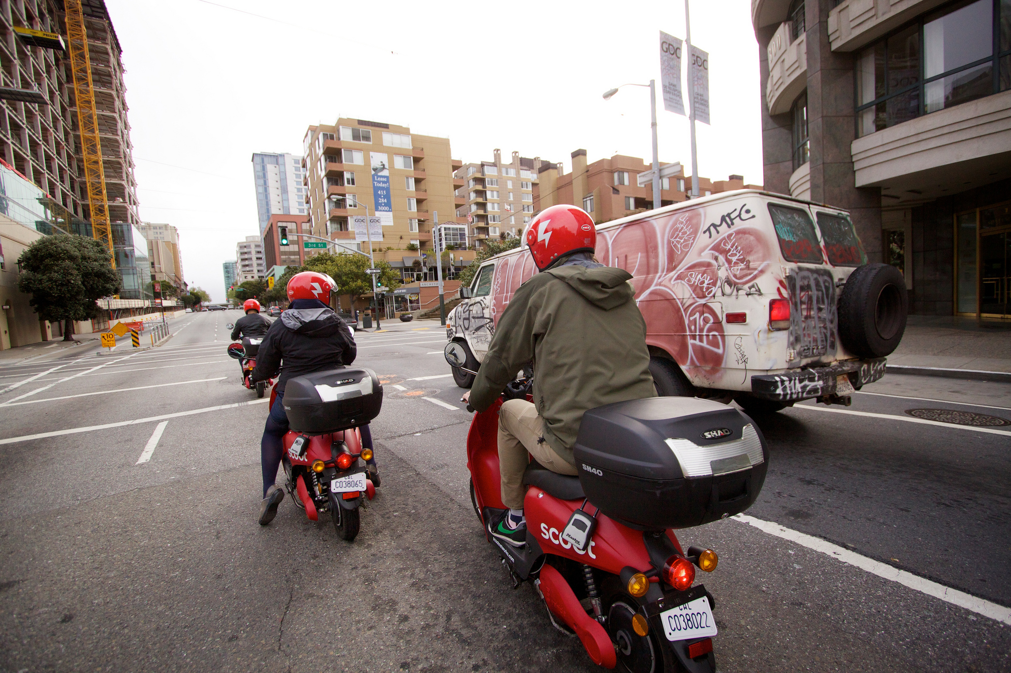 Two people in dark jackets ride red scooters down a busy city street in front of a white cargo van with red graffiti on the side.