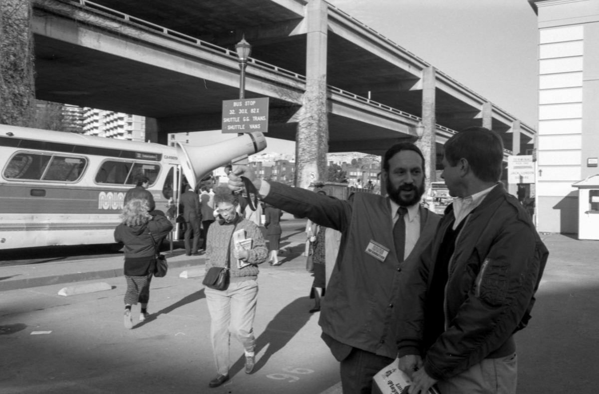 Bearded man in a suit and tie holding a megaphone speaks to another man in a windbreaker jacket with a Muni bus and other passengers in the background. The close Embarcadero Freeway is behind them.