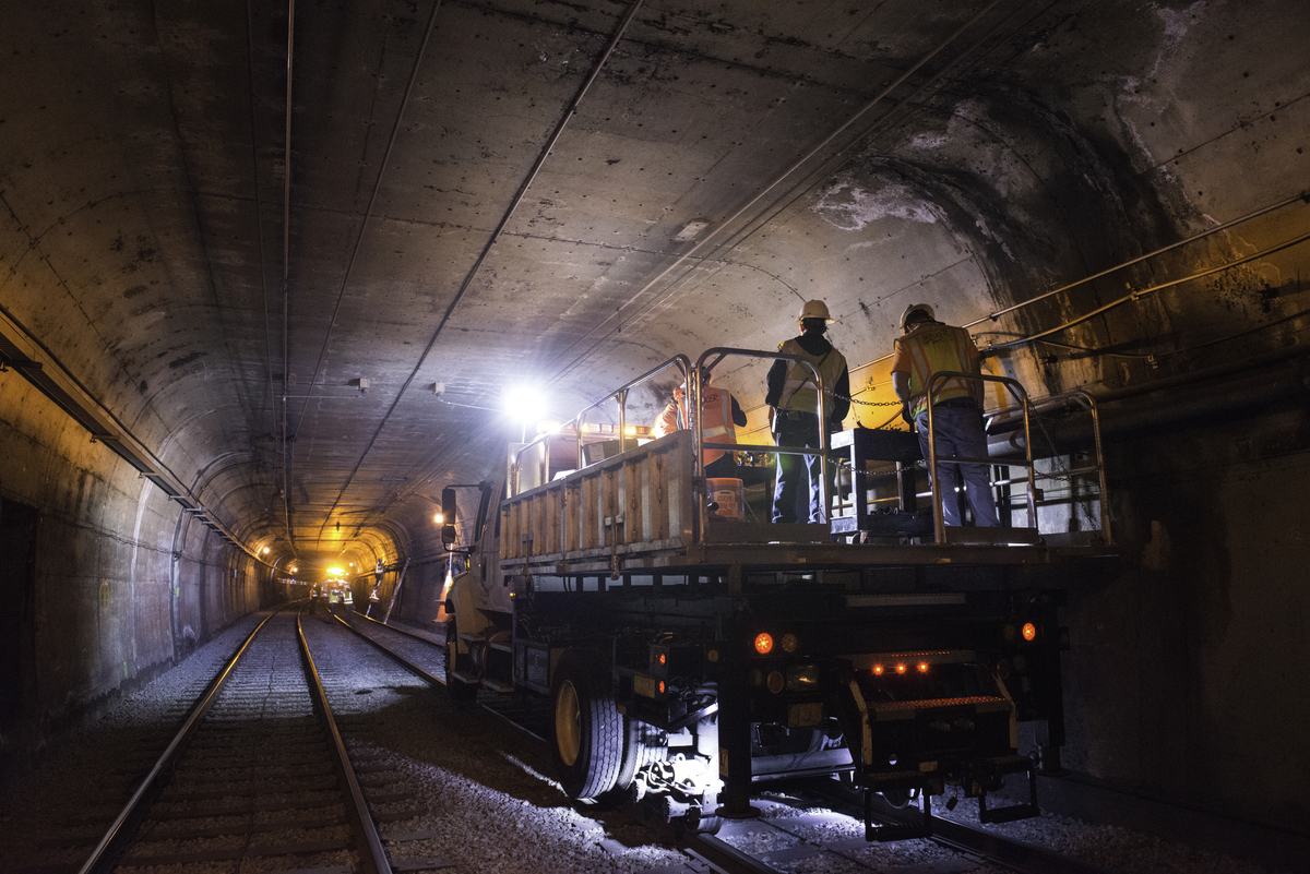 Construction crew in hard hats ride a special vehicle through the Muni subway tunnel with a welder's torch glowing in the background.