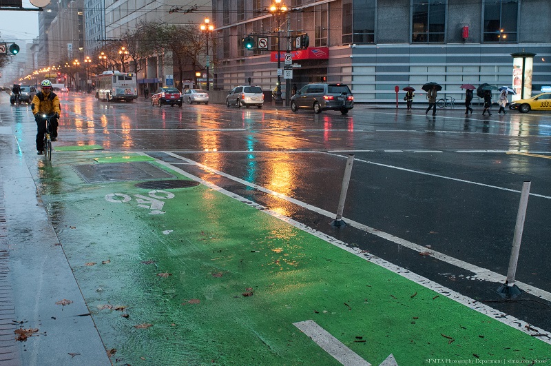 Looking east on Market Street on a wet day with the streetlights reflected in the green and black pavement, a bus and taxi traveling east and a bicyclist going west.