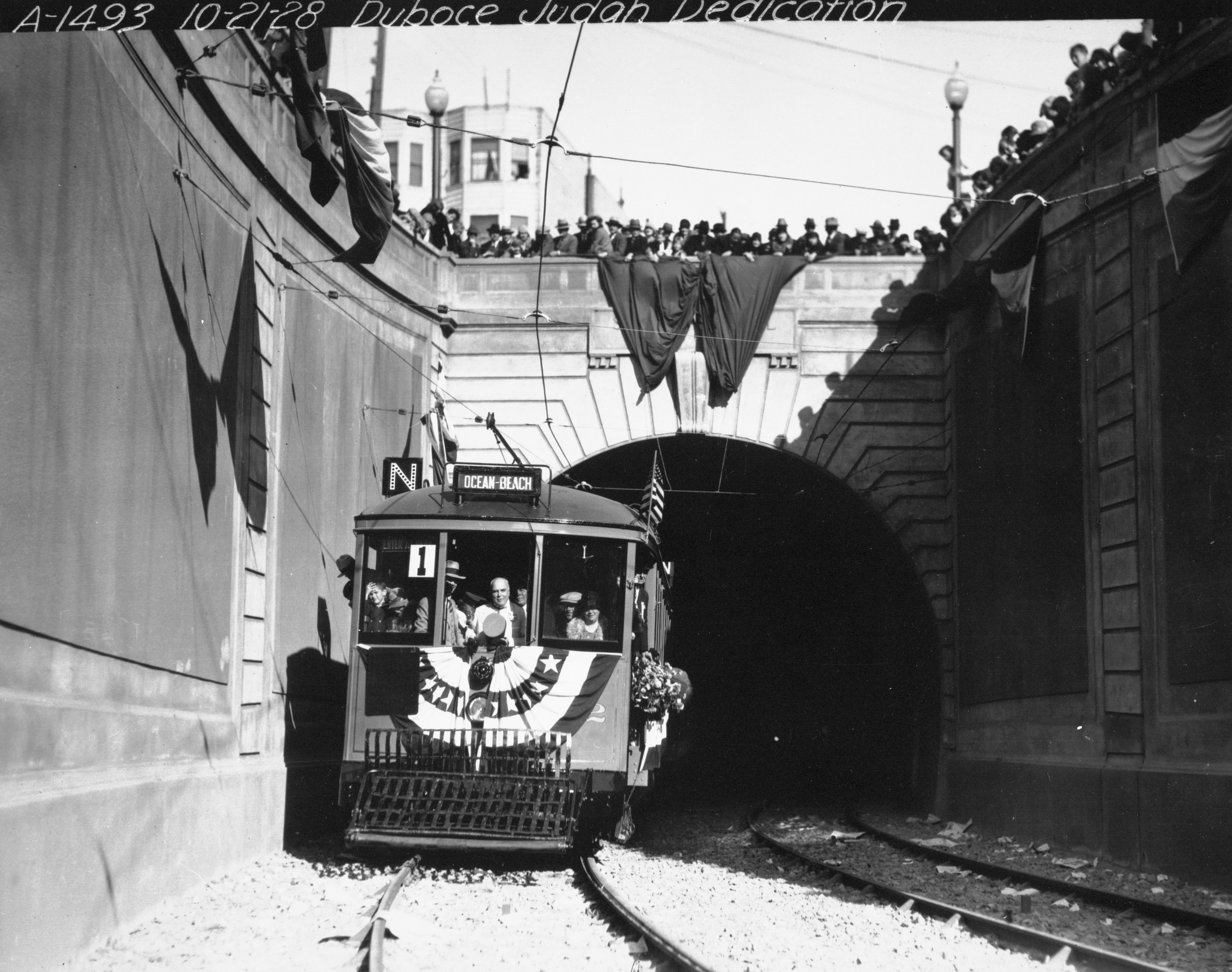 Black and white photograph from October 1928 opening of Sunset Tunnel. Streetcar exits tunnel on Duboce. Crowds and bunting circle the tunnel opening from the streets above.