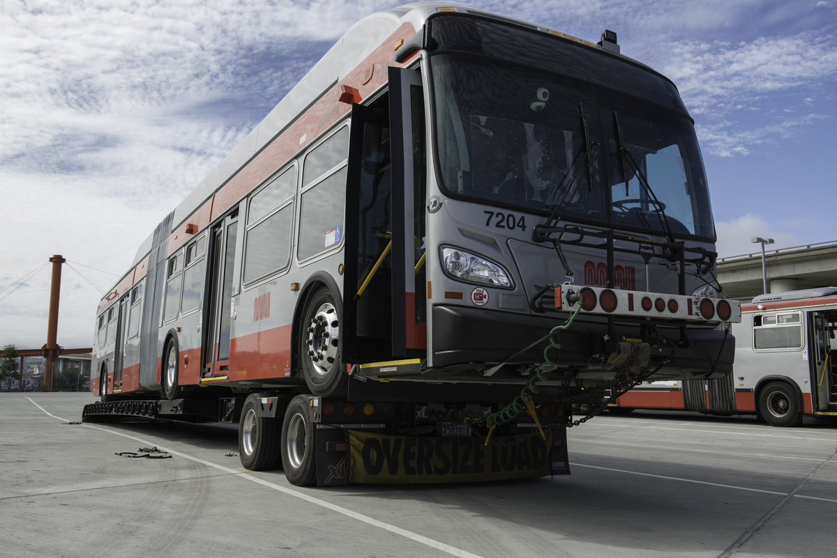 Low-angle photograph of a new gray and red Muni bus on a flatbed truck with the "oversized load" banner hanging below the truck and a cloud-speckled sky in the background.