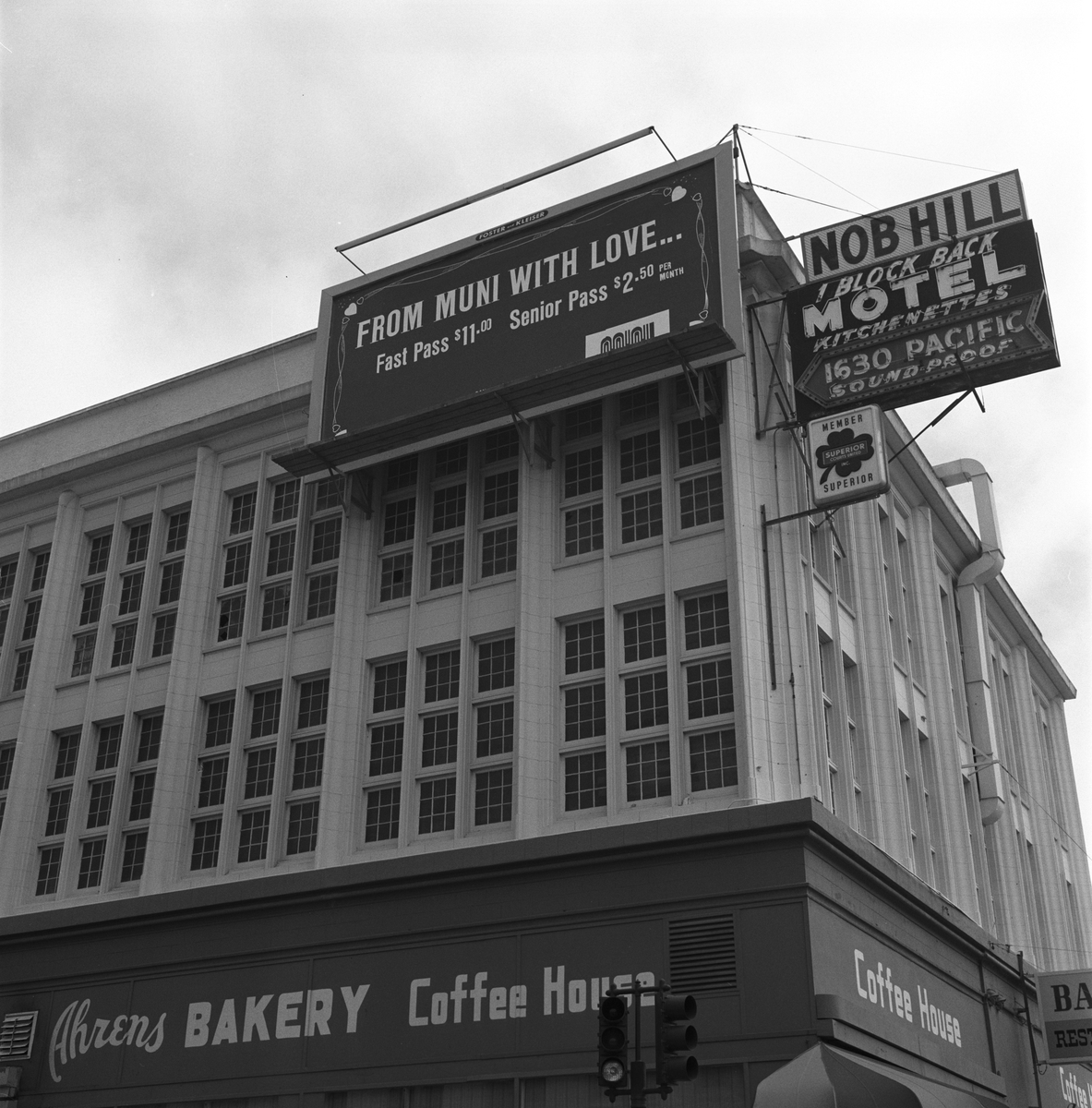 Black and white photo of a billboard and other signs on a large, square building. Billboard reads, "From Muni with Love..., Fast Pass $11.00, Senior Pass $2.50"