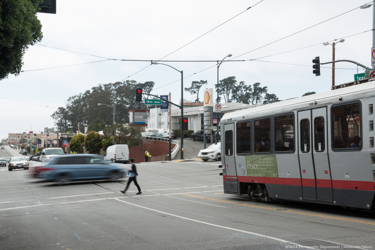 Busy traffic intersection with a light rail vehicle on the right, a pedestrian crossing and a blurred blue sedan going through the intersection.