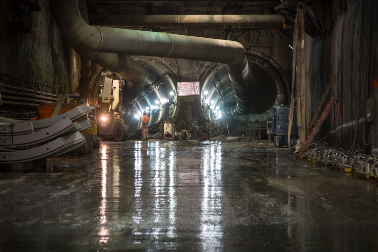 Northbound and southbound Central Subway tunnel entrances from the subterrenean launch box. Pipes and constrction equipment in the foreground, crews work at entrance of tunnel on the left.