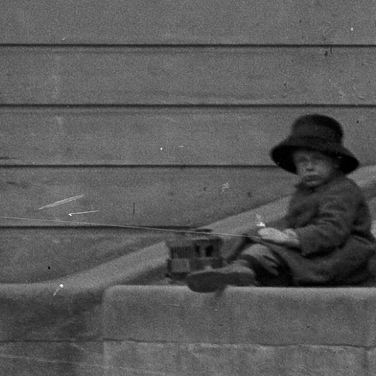 Detail view from Sacramento Street photo showing small boy sitting atop a low retaining wall wearing a floppy hat, overcoat and playing with a model streetcar toy.  February 3, 1913