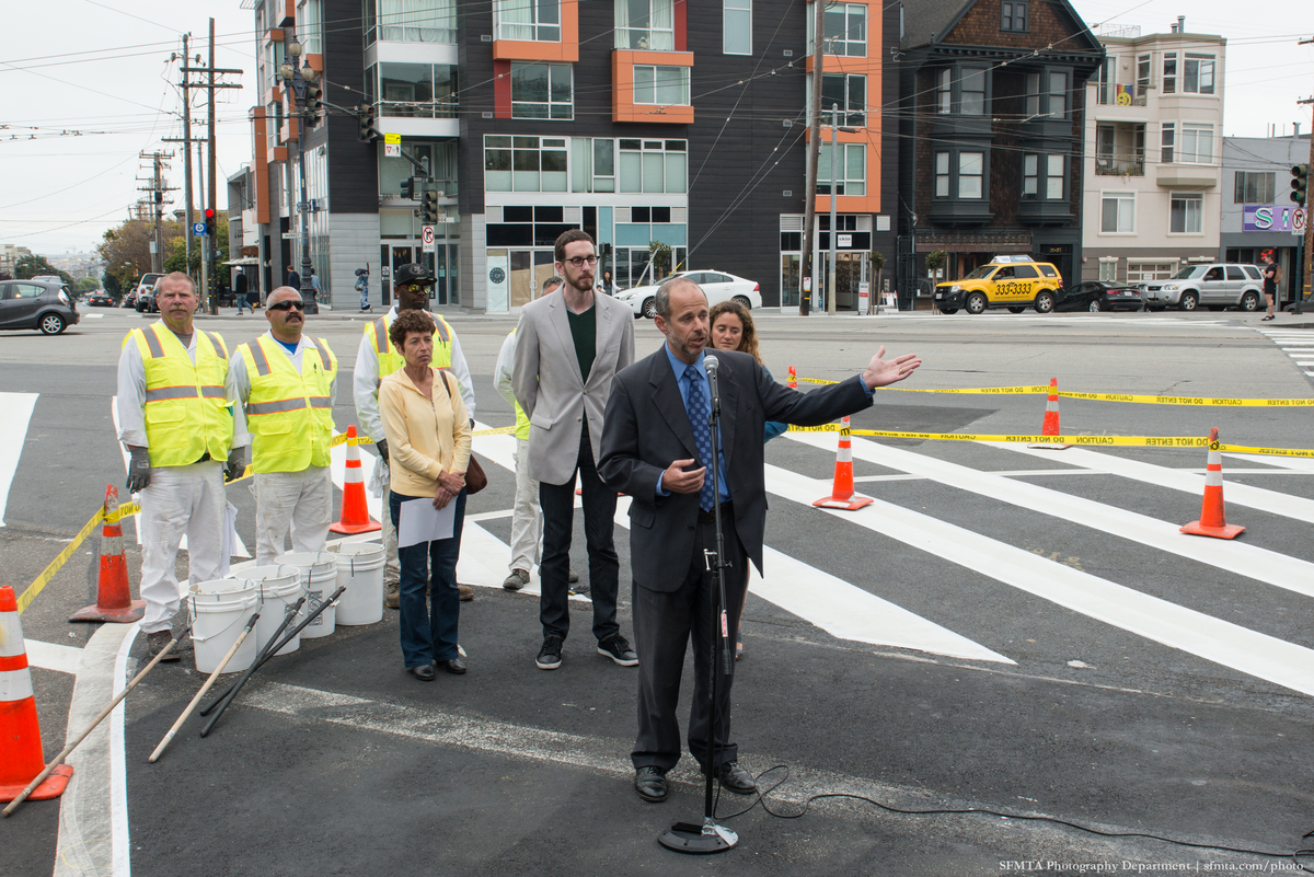 Ed Reiskin speaks at the microphone with Scott Wiener and two women standing behind him. The paint crew in white clothes and yellow safety vests stand behind them along Market Street.