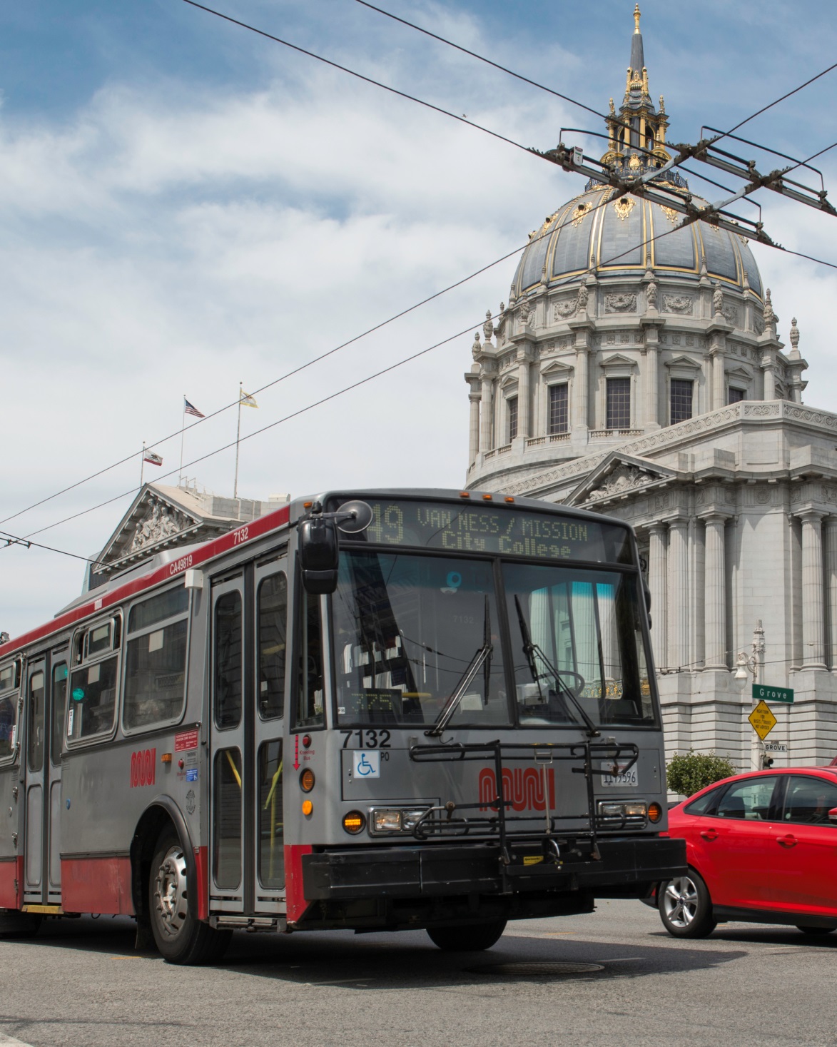A red and gray Muni bus travels next to a red car on Van Ness Avenue with City Hall and overhead wires in the background.