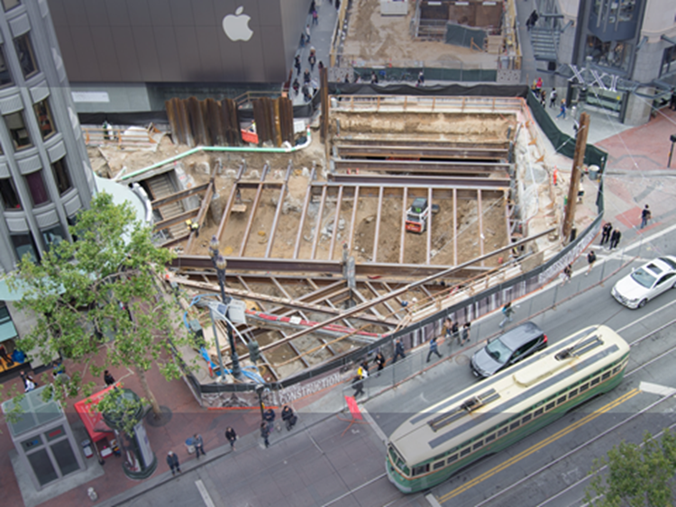 Aerial photograph of the subterranean construction site at Market, Stockton and Ellis streets with a streetcar, autos and pedestrisans passing by.