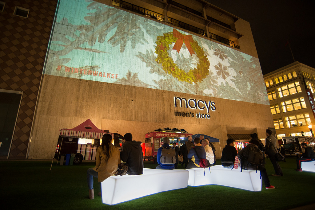 Visitors sit on white lighted benches at night in front of the Macy's store with the projected light show of a Christmas wreath and snowflakes on the building above them.