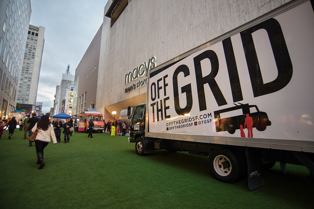 A truck sits on an astroturf-lined street next to Macy's Men's Store with "Off the Grid" in bold black text on the side.