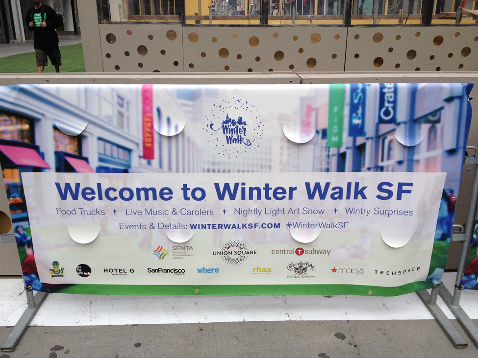 Event sign on the street, "Welcome to Winter Walk SF" with sponsor logos and website, "winterwalksf.com" and hashtag "#WinterWalkSF"