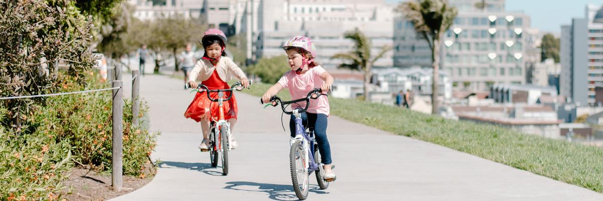 Two children biking in a park on a path with the San Francisco skyline behind