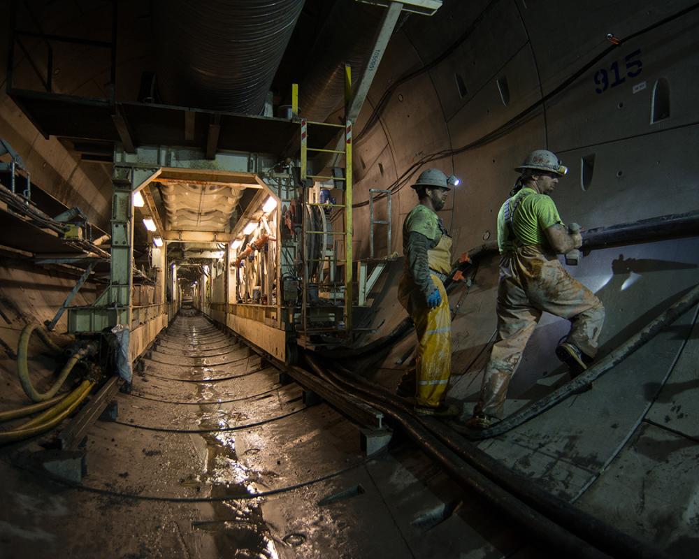 Workers attach new sections of utility pipes and conduit to the southbound tunnel wall as the trailing gear moves forward.