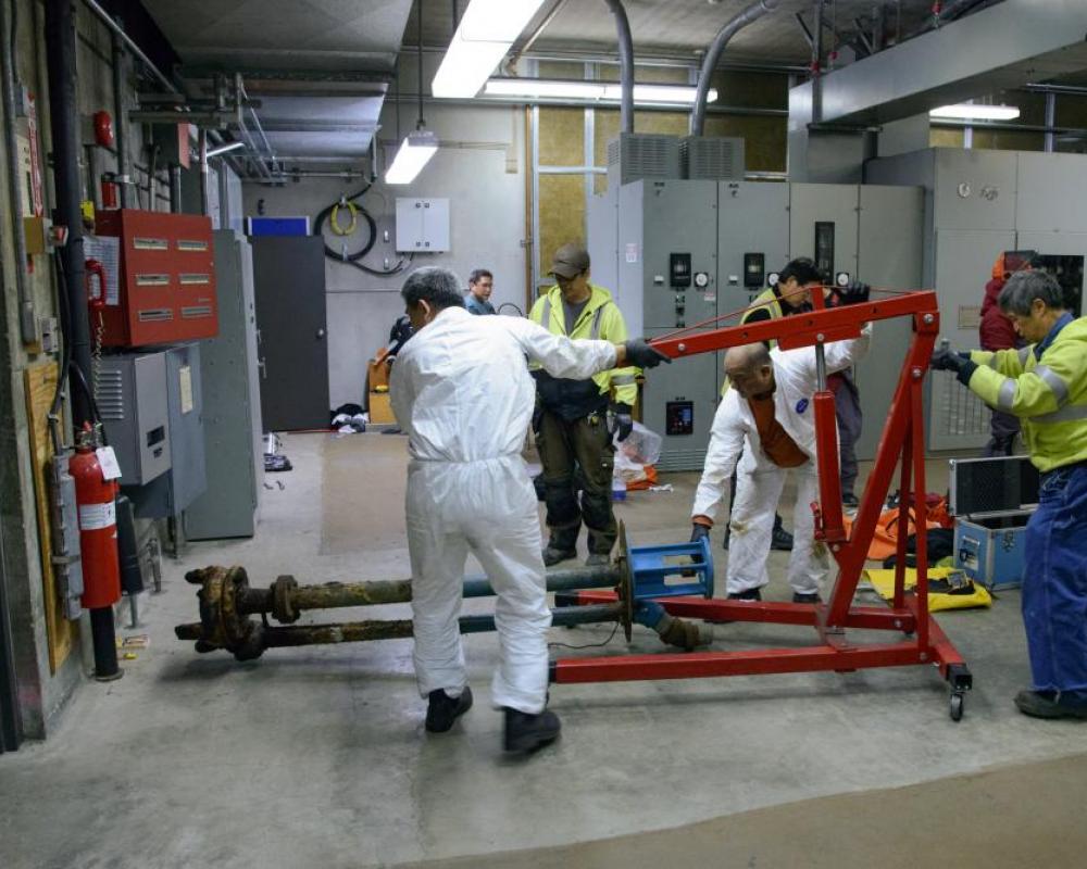 Several crew members holding a large device instrument inside a systems shop location. 