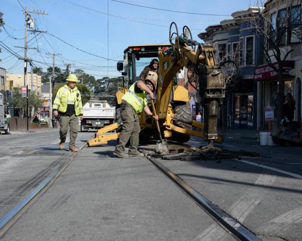 Crew member by the rail track and an excavator with a shovel along the street with houses lined along the side. 