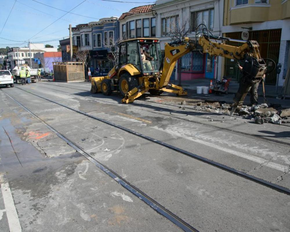 Excavator with extended arm and alongside the rail tracks and residential homes.