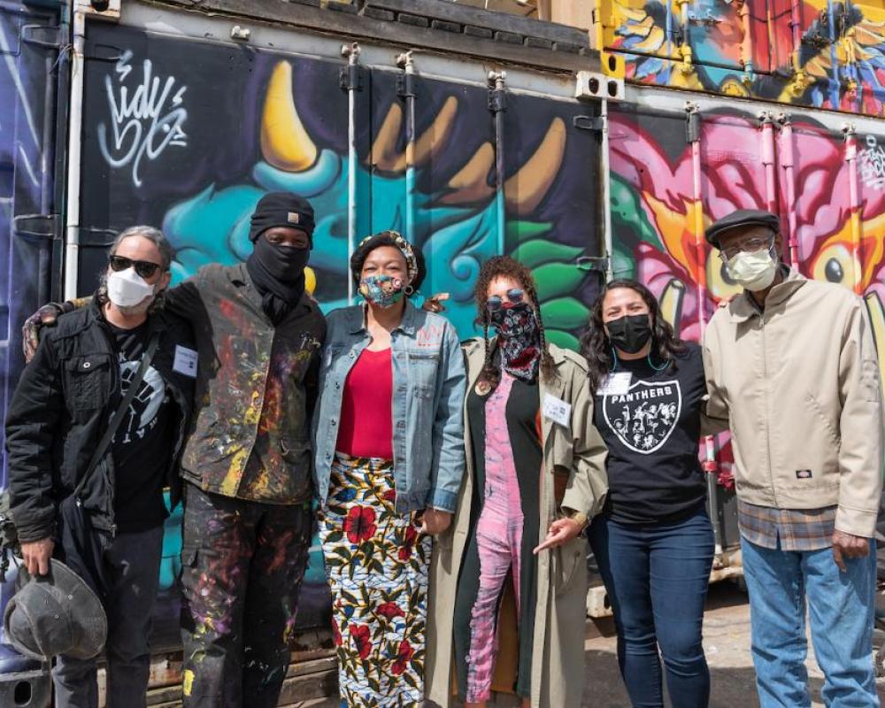 Several individuals with masks huddle for a group photo in front of a colorful mural backdrop in an our setting. 