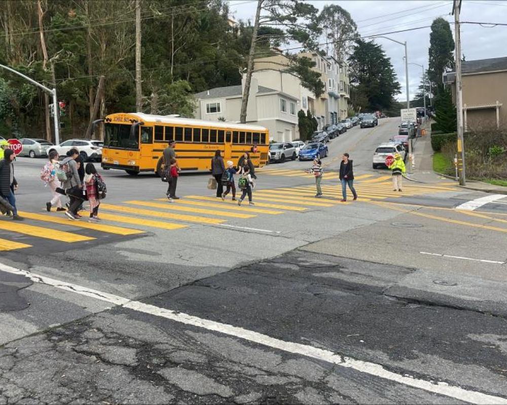 students using a crosswalk to cross clarendon avenue at panorama drive a bus is waiting in the intersection.