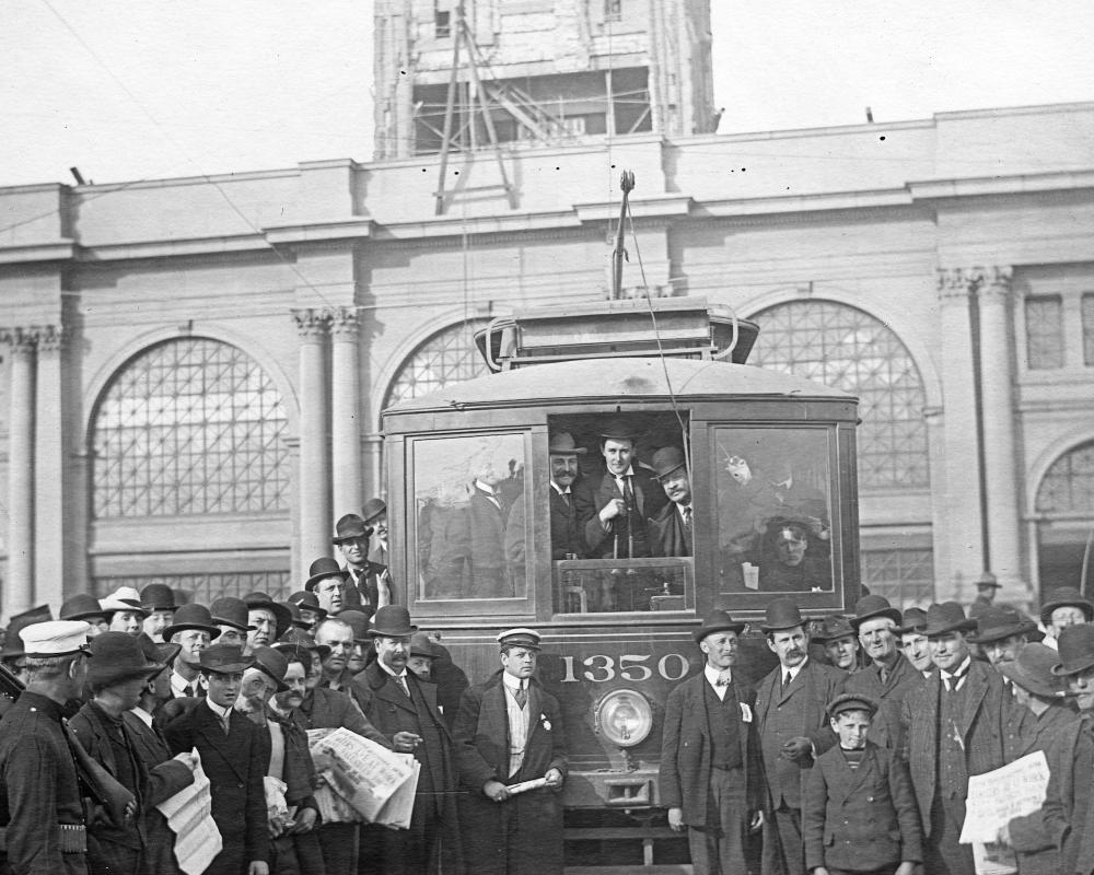 black and white photo of a crowd of people standing in front of a streetcar at the foot of the ferry building