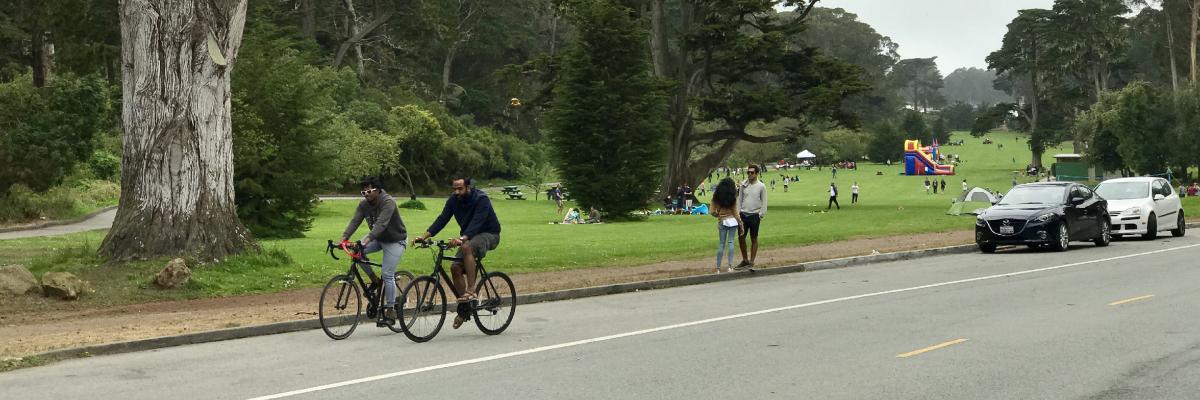 Image of people riding bicycles, a couple standing, and cars parked along John F. Kennedy Drive in Golden Gate Park.