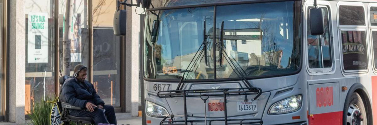 : Image of a Muni bus pulled to the curb with its wheelchair ramp deployed as a transit rider using a wheelchair rolls onto bus 