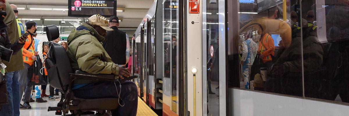 A person in a motorized wheelchair is wearing a tan hat and green jacket while waiting to board the T-line in the Central Subway