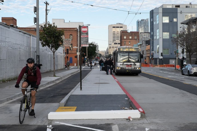 A man rides in a bike lane between the sidewalk and a concrete island where people board a Muni bus on 11th at Harrison.